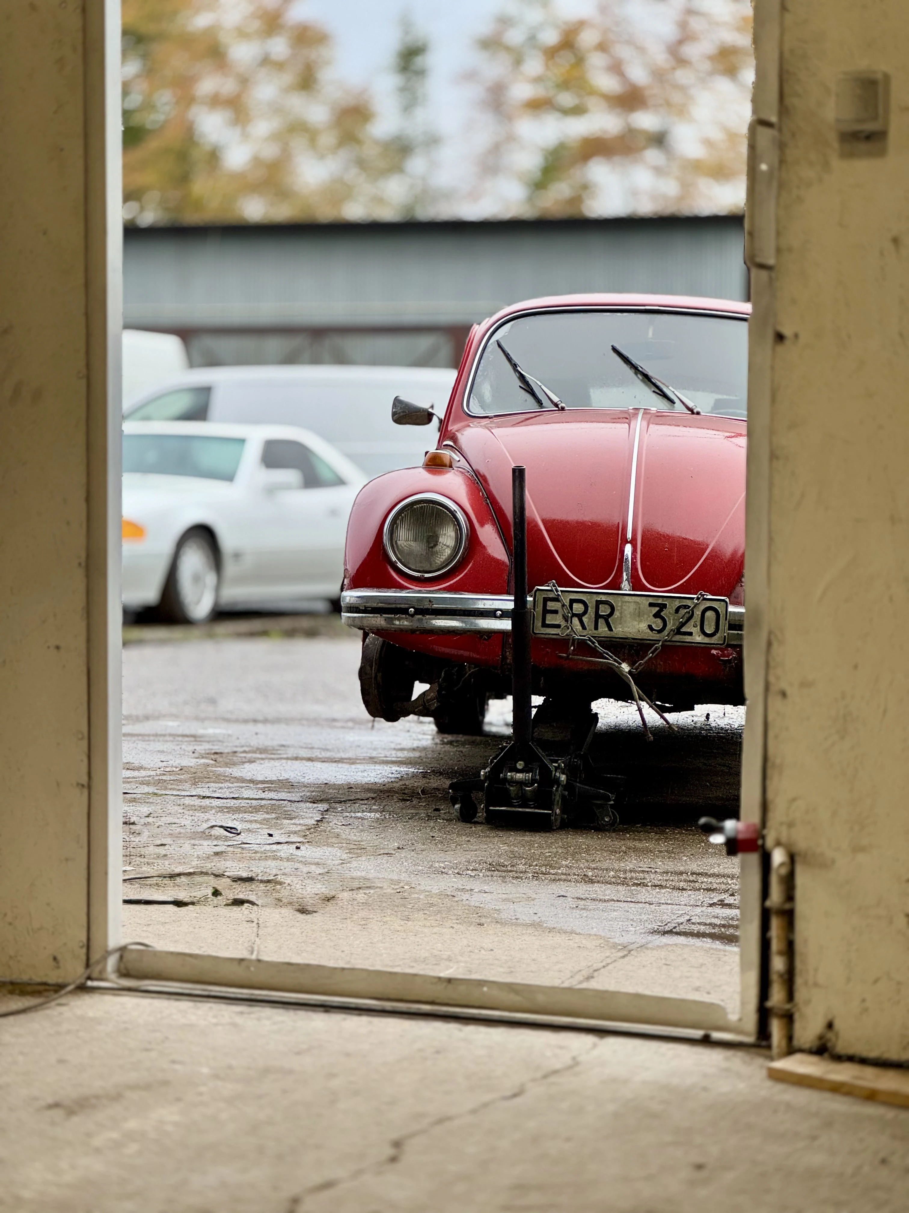 Czerwony Volkswagen Garbus stoi przed warsztatem z podniesionym przodem na lewarku; w tle widoczne inne samochody i jesienne drzewa.