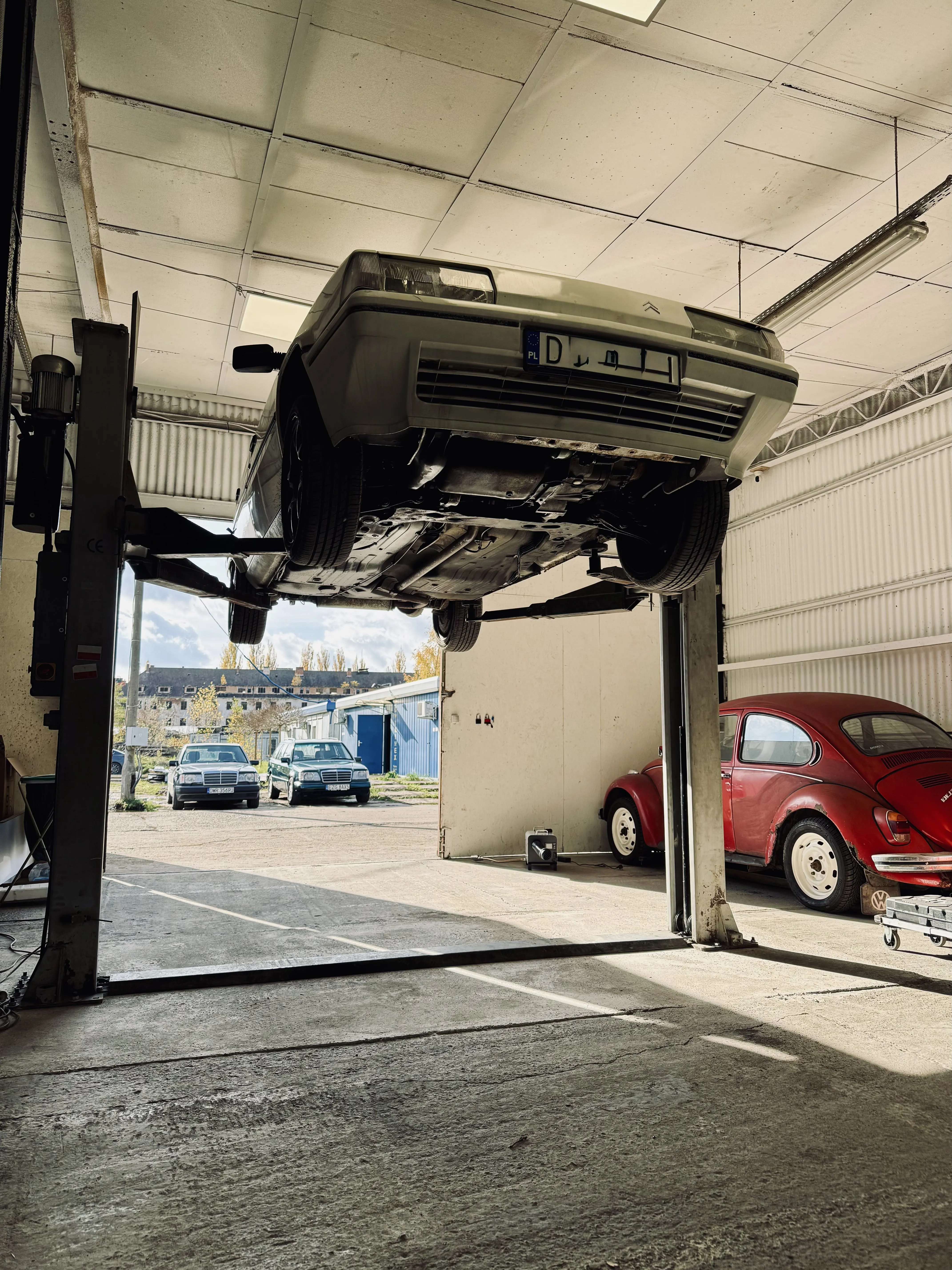 Samochód osobowy uniesiony na podnośniku w warsztacie samochodowym, z widocznym podwoziem; w tle widać czerwonego Volkswagena Garbusa i inne auta na zewnątrz.