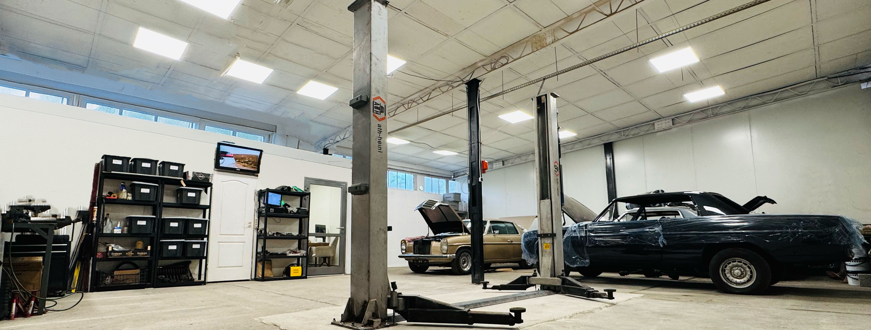 Wnętrze warsztatu samochodowego z podnośnikiem i dwoma klasycznymi samochodami w trakcie naprawy.